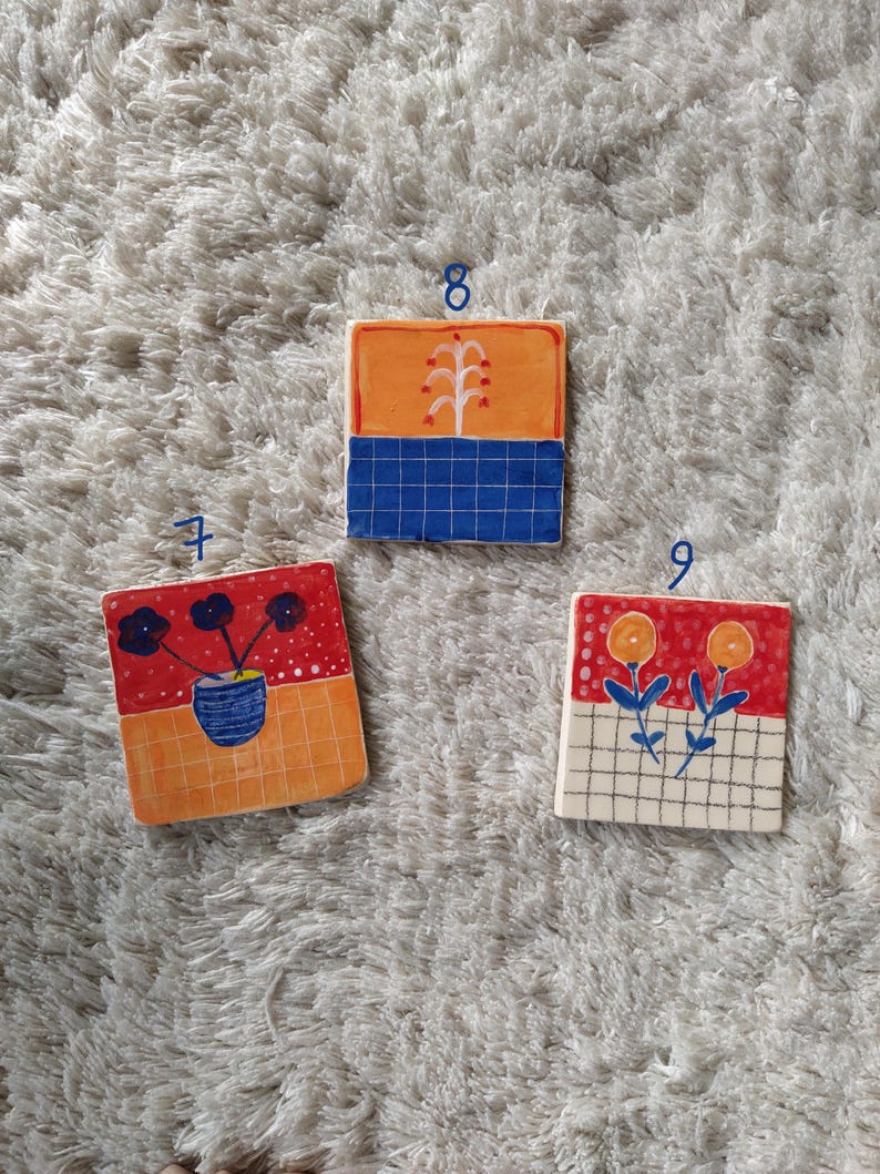 May include: Three square ceramic tiles with different floral designs. The first tile has a red background with black flowers in a blue vase. The second tile has an orange background with a white tree and blue grid. The third tile has a red background with yellow flowers and a white grid.