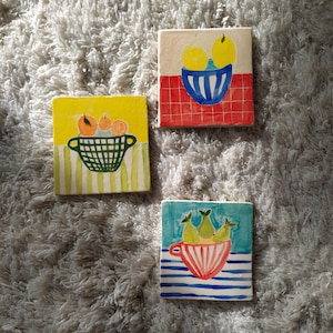 May include: Three ceramic tiles with hand-painted fruit designs. The first tile features a yellow bowl with three lemons on a red and white checkered background. The second tile features a green basket with three oranges on a yellow background with green and white stripes. The third tile features a red and white striped bowl with three green pears on a blue background with white stripes.