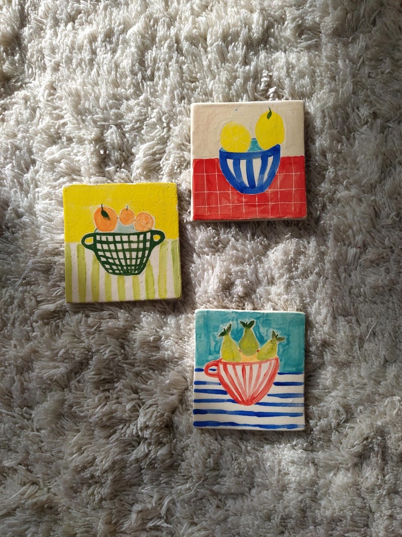 May include: Three ceramic tiles with hand-painted fruit designs. The first tile features a yellow bowl with three lemons on a red and white checkered background. The second tile features a green basket with three oranges on a yellow background with green and white stripes. The third tile features a red and white striped bowl with three green pears on a blue background with white stripes.