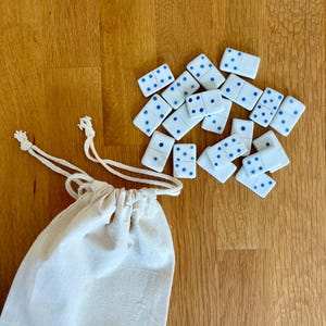 May include: A set of white dominoes with blue dots scattered on a wooden surface next to a small, off-white drawstring bag. The dominoes are rectangular and feature various combinations of dots. The bag is made of a light-colored fabric.