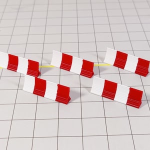 May include: Five miniature red and white barricades are arranged on a grid. The barricades have a rectangular shape with alternating red and white stripes. The background is a white grid with a yellow line.