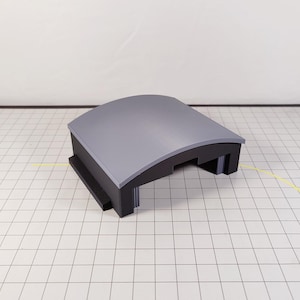 以下が含まれることがあります： 曲がった屋根を持つグレーと黒の建築模型。 模型は長方形のベースと丸みを帯びた上部を持ち、モダンなデザインを示唆しています。 オブジェクトはグリッドの背景に配置されています。