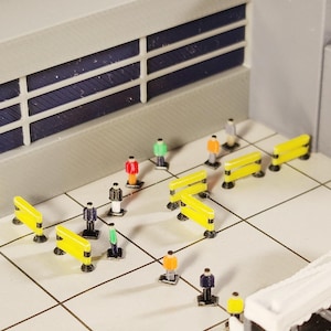 Könnte beinhalten: Miniatur-Flughafenszene mit kleinen Figuren und gelben Absperrungen. Die Szene umfasst ein Gebäude mit grauer Fassade und einem Fenstergitter mit dunkelblauen Innenräumen. Die Figuren sind in verschiedenen Farben gekleidet, was auf eine geschäftige Flughafenatmosphäre hindeutet.
