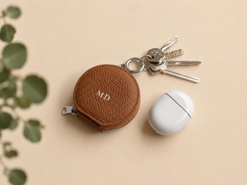 Keychain zip pouch in brown leather with initials MD shown next Apple Airpods.
