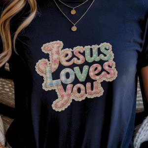 May include: Navy blue t-shirt featuring the text "Jesus Loves You" in a colourful, floral design. The lettering is in a patchwork style with pink, blue, and green colours. A gold necklace is visible.