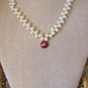 Può includere: Una collana di perle con un pendente rosso. La collana è composta da numerose piccole perle bianche. Il pendente è una pietra rossa rotonda con una piccola gemma trasparente al centro. La collana è esposta su un manichino.