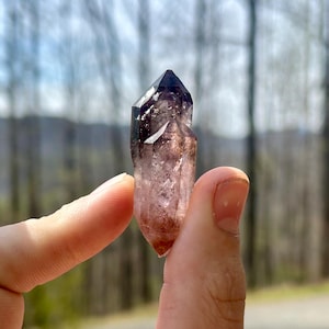 May include: A natural crystal with a dark purple tip and a translucent, light purple body. The crystal is held between two fingers, showcasing its pointed shape and natural texture. The background is blurred, with trees and a hint of a road visible.