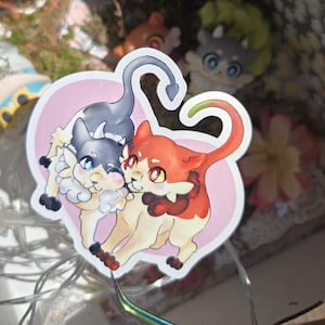 May include: A sticker featuring two cartoonish cat-like creatures. One is gray and white, the other is orange and cream. They are set against a pink heart-shaped background. The sticker has a white border and is likely for decorative purposes.