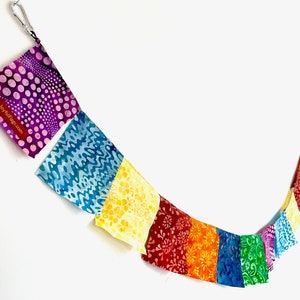 May include: A string of multi-colored fabric flags featuring various patterns including dots, waves, and floral motifs. The flags are arranged in a spectrum of purple, blue, yellow, red, orange, and green. A red tag with the text JoyfulFlags.com is attached.