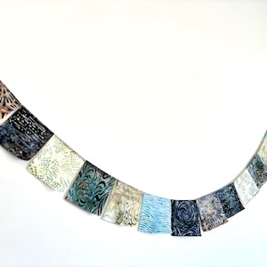 May include: A decorative bunting banner featuring a series of square fabric flags in various shades of blue, teal, and cream. Each flag displays unique batik-style patterns, including swirls, dots, and geometric lines, suspended on a string.