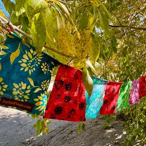 May include: A string of colorful fabric flags hangs from a tree branch. The flags feature various patterns, including floral designs and ladybugs. The first flag is teal with yellow floral patterns, and the text "joyfulflags.com".