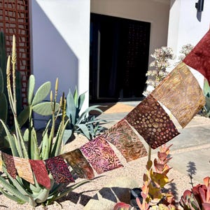 Pode incluir: Uma faixa de tecido colorida com uma variedade de padrões e cores, incluindo marrom, vermelho e rosa. A faixa está pendurada entre duas plantas em um jardim.