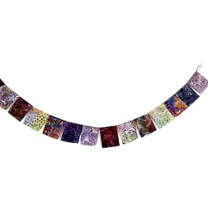 Puede incluir: Un banderín de tela colorido con una variedad de patrones florales en tonos de morado, rosa y verde. El banderín está colgado en una pared blanca.