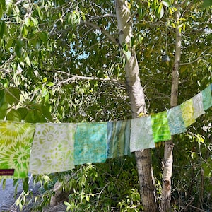 May include: A string of colorful fabric squares, each with unique patterns and shades of green, blue, and white, hangs from a tree branch. The fabric squares are printed with various designs, including tie-dye and floral patterns. The website joyfulflags.com is visible on one of the squares.