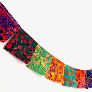 May include: A string of rectangular fabric flags hanging in a curve, featuring vibrant batik-style floral patterns in shades of purple, pink, green, orange, and teal. The largest flag on the left displays the text JoyfulFlags.com on a small label.