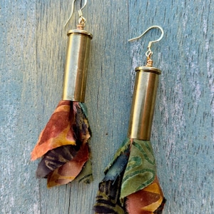 May include: A pair of dangle earrings featuring polished brass bullet casings as the main body. Each earring is finished with a gold-toned wire hook and a decorative tassel made from layered, frayed fabric scraps in shades of orange, green, and brown.