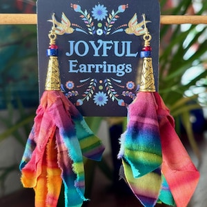 May include: Joyful earrings with a gold-colored cone-shaped base and colorful fabric tassels. The earrings have a blue, red, and gold bead accent. The earrings are displayed on a navy blue card with the words "JOYFUL Earrings".