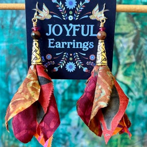 May include: A pair of dangle earrings with a fabric tassel in shades of red, orange, and brown. The tassel is attached to a gold-colored metal bead and a gold-colored metal hook. The earrings are labeled "Joyful Earrings".