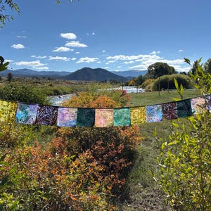 Peut inclure: Une guirlande horizontale de drapeaux en tissu rectangulaires présentant une variété de motifs colorés, notamment des rayons de soleil jaunes, des teintures bleu et violet, des textures vert foncé et des motifs géométriques complexes.