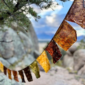 Puede incluir: Una guirnalda de banderines triangulares de tela con una variedad de patrones inspirados en el otoño en tonos oro, naranja, marrón y rojo intenso. Los banderines están conectados por un cordel fino y muestran texturas intrincadas y diseños.