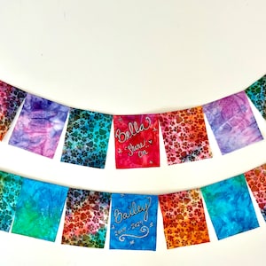 May include: Two colorful fabric banners with paw print patterns and the names "Bella" and "Bailey" written on them. The banners have a variety of colors, including red, blue, and purple. Each banner has a silver carabiner clip.