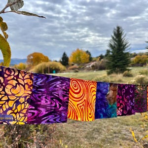 May include: A string of colorful fabric flags with various patterns, including floral and abstract designs. The flags are in shades of purple, orange, yellow, and blue. The text "joyfulflags.com" is visible on one of the flags. The flags are strung across a natural outdoor setting.