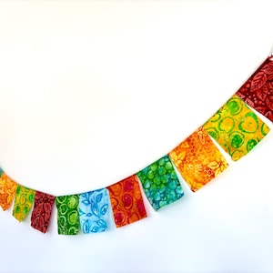 May include: A colorful fabric banner with a rainbow of colors, including red, orange, yellow, green, blue, and purple. The banner is made of fabric squares with different patterns and is strung together with a white cord.