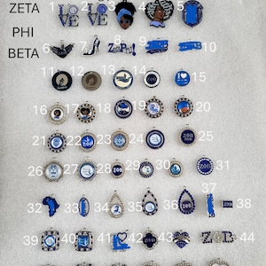 May include: A collection of blue and silver pendants with various designs, including hearts, shields, and circular charms. Some pendants feature the letters ZETA, PHI, and BETA, along with the text "LOVE" and "ZΦB". The image displays a variety of styles.