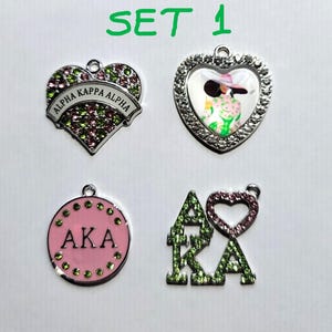 May include: Set of four charm pendants featuring the Greek letters AKA and Alpha Kappa Alpha. The charms are silver-toned and decorated with pink and green glitter. One charm is a heart with the letters AKA inside, another is a circle with the letters AKA inside, another is a heart with the words Alpha Kappa Alpha inside, and the last is a heart with a picture of a woman inside.
