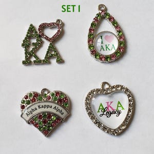May include: Four decorative charms with the letters AKA and heart shapes. The charms are embellished with green, pink, and clear rhinestones. One charm has the text "I ❤️ AKA", and another says "Alpha Kappa Alpha".