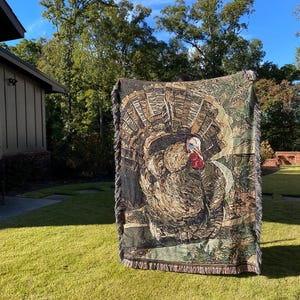 Puede incluir: Una manta de tejido de tapicería que presenta una imagen detallada de un pavo salvaje con una combinación de colores marrón, gris y verde. La manta tiene bordes con flecos.