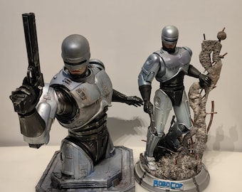 May include: Two Robocop statues. One is a bust holding a gun, the other is a full figure. Both are silver and black with detailed armor. The full figure stands on a base with the word "ROBOCOP".