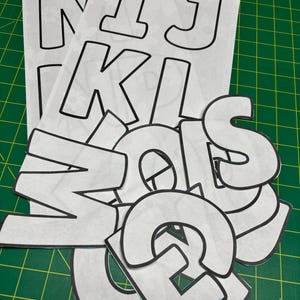 Peut inclure: Découpes de lettres blanches épelant "NIKI" et "MOLES". Les lettres ont un contour noir et sont éparpillées sur un tapis de coupe vert. Les lettres sont destinées à l'artisanat ou au scrapbooking.