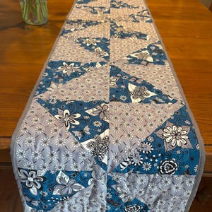 Puede incluir: Camino de mesa acolchado rectangular con un estampado floral en tonos azules, grises y blancos. El diseño de patchwork presenta varios motivos florales. Los bordes están acabados con un borde gris.