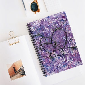 Puede incluir: Cuaderno de acuarela morado y azul con un diseño de corazón de tinta negra. El cuaderno tiene una encuadernación en espiral.