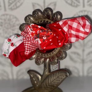 May include: A decorative metal flower-shaped holder with red fabric scrunchies. The scrunchies feature patterns like cherries, polka dots, and gingham. The holder is on a white surface.