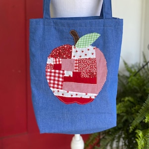 May include: Blue denim tote bag with a large apple applique. The apple is made of red and white patterned fabric pieces, including gingham and floral prints. The bag has two shoulder straps and a simple rectangular shape.