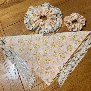 May include: A set of floral patterned accessories, including two scrunchies and a bandana. The scrunchies and bandana feature a pink background with yellow floral designs and white lace trim. The items are arranged on a wooden surface.