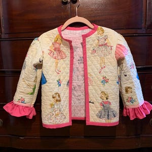 May include: A quilted jacket with a cream-colored exterior featuring a repeating pattern of vintage doll illustrations in various dresses. The jacket has pink trim, ruffled cuffs, and a pink lining.
