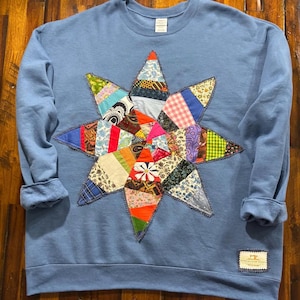 May include: A dusty blue crewneck sweatshirt featuring a large, multi-pointed star applique on the front. The star is constructed from various fabric scraps in diverse colors, patterns, and textures, outlined with dark stitching. A small label is at the hem.