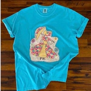 May include: A turquoise t-shirt with a crew neck and rolled-up sleeves. The shirt features a colorful, appliqued design of a figure in a dress, holding a parasol. The design includes floral and star accents.