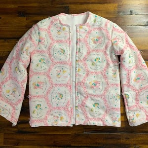 May include: A quilted jacket with a white background and a repeating pattern of pink circles with illustrations of children. The jacket has long sleeves and a white lining. The edges of the jacket are trimmed with a frayed, textured material.