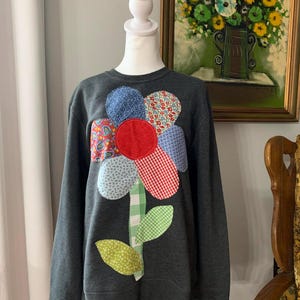 May include: Dark gray sweatshirt featuring a large, colorful flower applique. The flower has a red center, blue, red, and patterned petals, and a green and white checkered stem and leaves. The sweatshirt is displayed on a mannequin.