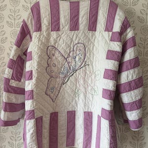 May include: A quilted jacket featuring a white and lavender striped pattern on the sleeves and borders. The center panel displays a diamond-stitched texture with an embroidered butterfly and floral design in shades of purple, pink, and green.