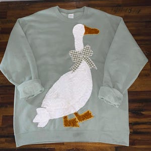 May include: A light green sweatshirt featuring a large white goose applique with a textured body, brown beak and feet, and a plaid bow. The long-sleeved top has ribbed cuffs and a crew neck.
