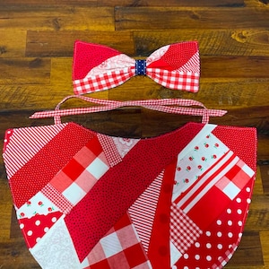 May include: A red and white patchwork bib featuring various patterns including gingham, polka dots, and cherry prints, accompanied by a matching bow tie with a dark blue center. The bib has fabric ties at the top for securing around the neck.