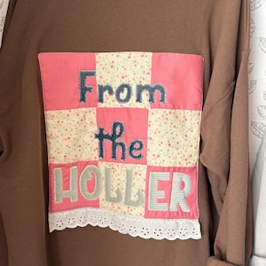 May include: A brown crewneck sweatshirt featuring a large, square, patchwork-style appliqué on the back. The appliqué consists of pink and floral-patterned fabric squares with the embroidered text "From the HOLLER" and white lace trim.