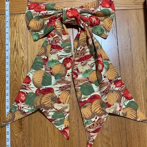 May include: A large fabric bow featuring a colorful autumn harvest pattern. The bow is adorned with images of pumpkins, apples, and corn on a cream background. The bow's width is approximately 20 inches.