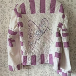 May include: A quilted jacket featuring a white base with lavender striped panels on the sleeves and borders. The back panel displays a large embroidered butterfly design in lavender and blue thread, set against a diamond-stitched quilted fabric texture.