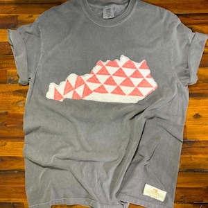 May include: A charcoal gray short-sleeve t-shirt featuring a central applique in the shape of the state of Kentucky. The applique has a white and coral pink geometric triangle pattern with a textured, frayed edge. A small rectangular label is at the hem.